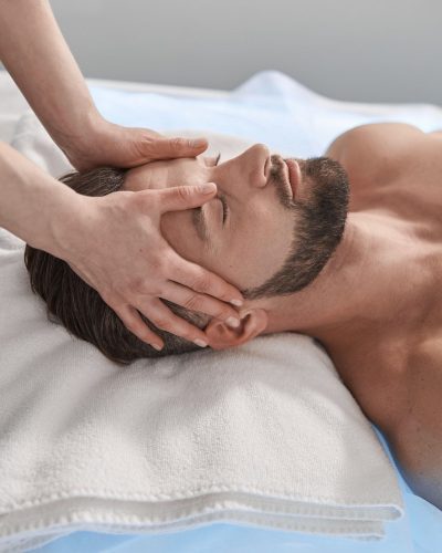 Massage therapist works with head of male lient lying on couch in contemporary clinic